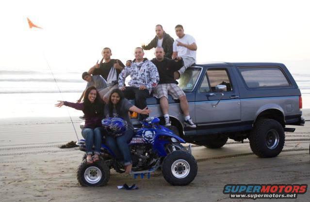 1987 Ford Bronco with people sitting on it and an ATV in front on a beach