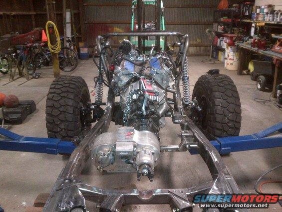Custom off-road vehicle frame with engine and transmission installed, sitting on lift arms in a garage