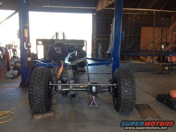 Jeep CJ frame with large tires, being worked on by a person with a grinder
