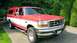 1993 Ford F-150 - This red and white Ford F-150 truck, equipped with a camper shell, is parked on a paved surface with greenery in the background.