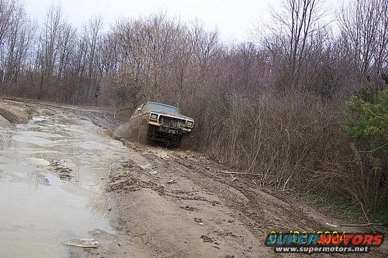 Brown 1978 Ford Bronco driving through mud on a dirt road, front 3/4 view