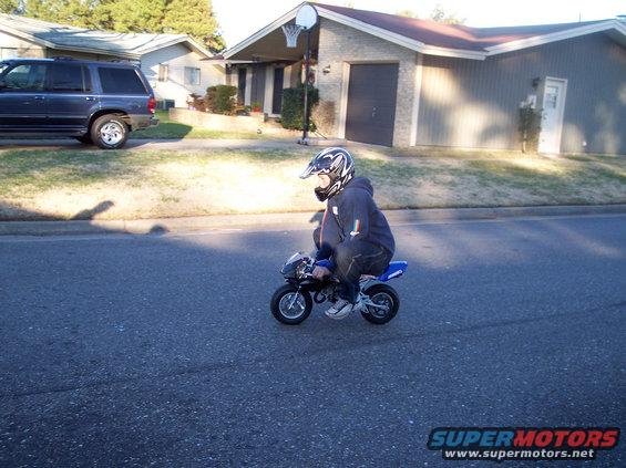 Person wearing a helmet rides a small blue and black pocket bike down a residential street. A dark SUV is parked on the left