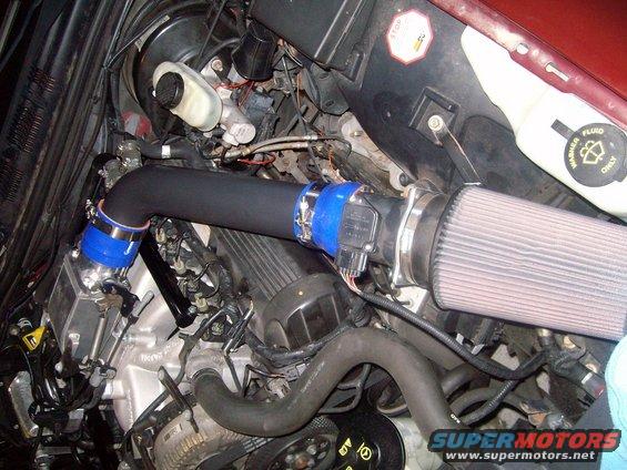 Engine bay with black intake tube, blue couplers, and a cone air filter. Some text is visible on a fluid reservoir and a stop sign
