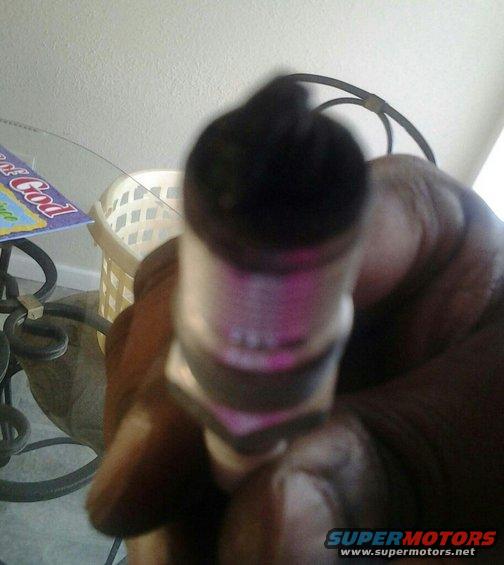 Spark plug held in hand, with a Supermotors.net logo visible at the bottom right