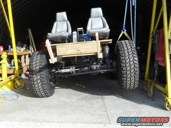 Custom off-road chassis with two bucket seats, large tires, and suspension components, suspended by chains and yellow cranes