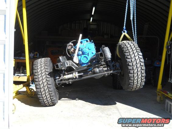 Chassis with blue engine being lifted by chains and yellow gantry