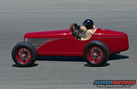 Red vintage race car with wire wheels and a driver wearing a helmet and racing suit on a paved track