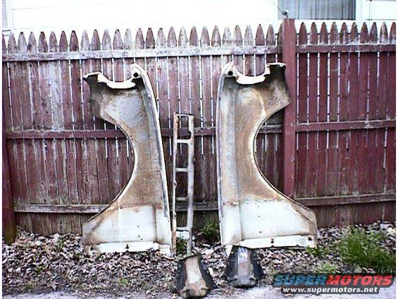 Two white car fenders and a metal trim piece sit on the ground in front of a wooden fence