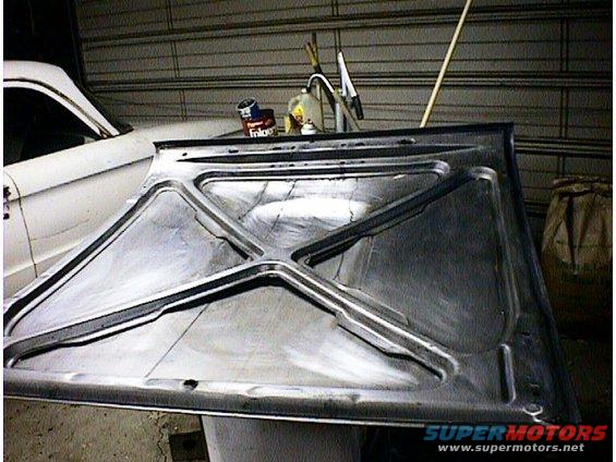Underside of a car hood, showing structural bracing. A white car is partially visible to the left