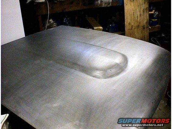 Car hood with a raised scoop, appears to be bare metal. A watermark says SUPERMOTORS