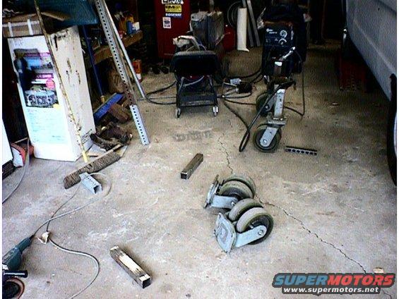 Garage interior with a workbench, tools, metal parts, and casters on the floor. Supermotors.net logo in the corner
