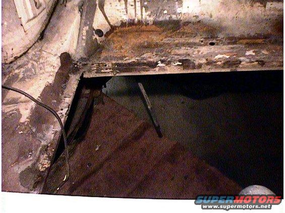 rusted car floor pan with a large hole revealing darkness below, wires visible