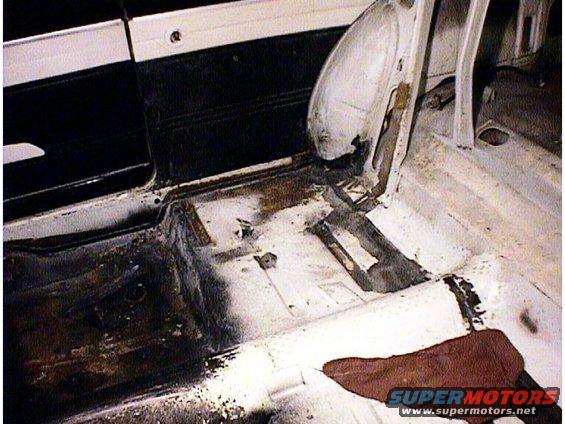 Interior floor pan of a car showing rust damage and repair work. Black door panel visible on left
