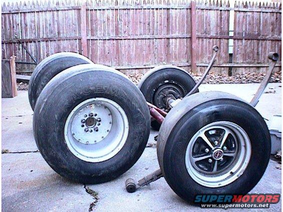 Four drag racing tires with white and silver rims, attached to suspension parts, resting on concrete