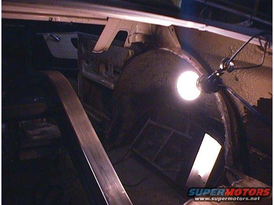 Interior view of a car's rear wheel well area, with a work light illuminating the space. Some structural components and a door panel are visible