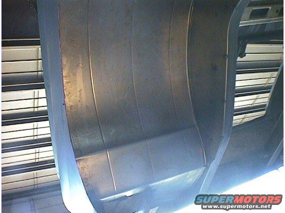 Underside of a car, showing metal panels and some structural elements. A watermark reads 'SUPERMOTORS.NET'