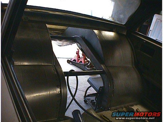 Interior view of a car with a roll cage installed, showing the rear seats and a floor jack in the background