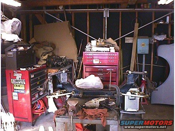 Car subframe assembly on a workbench in a garage, with toolboxes and various parts around