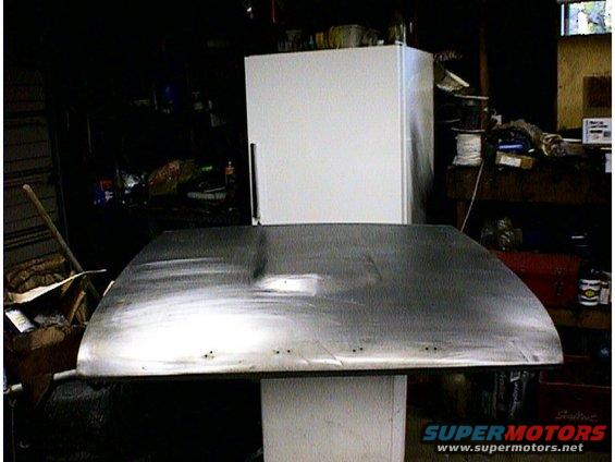Bare metal car hood on a stand in a garage, with a white refrigerator behind it