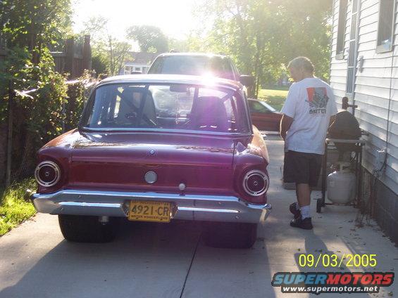 Rear view of a red 1960 Ford Falcon with a roll cage, parked outside next to a grill