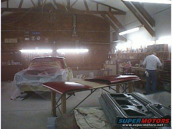 car covered in plastic sheeting in a workshop, with hood and door parts on tables