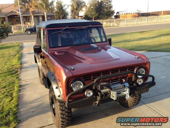 1968 Ford Bronco, red, with aftermarket hood scoop, winch, and soft top. Another blue SUV is parked in the background