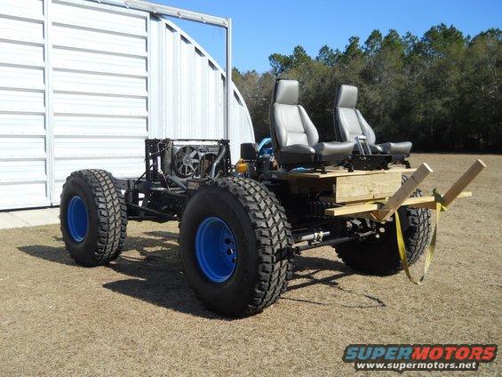Custom off-road vehicle chassis with two grey seats, large tires with blue rims, and exposed engine components