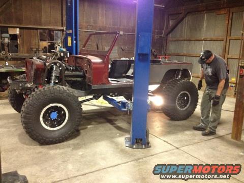 Red and grey Jeep CJ-series chassis on a lift, front wheels visible, man inspecting rear wheel