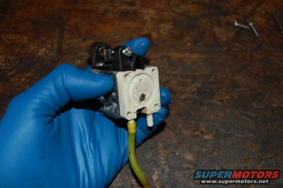Hand in blue glove holding a carburetor with yellow fuel line