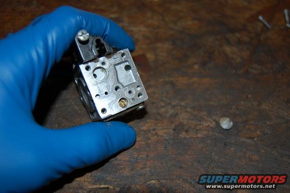 Gloved hand holding a metal carburetor component with small screws and holes