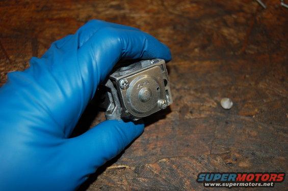 Hand in blue glove holds a small metal carburetor part with 'ZAMA' visible on it