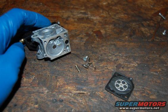 Carburetor disassembled on workbench, with blue-gloved hand holding main body