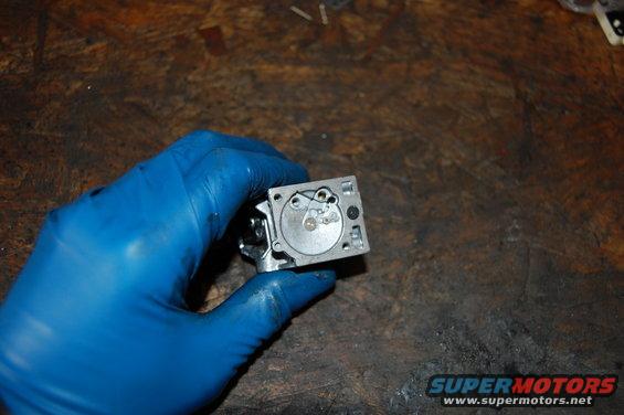 Carburetor part held in blue gloved hand over wooden surface