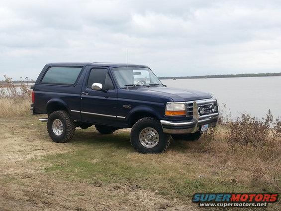 Blue 1995 Ford Bronco parked on grassy shore next to a large body of water