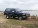 1995 Ford Bronco - A blue Ford Bronco is parked on a grassy area near a large body of water under a cloudy sky.