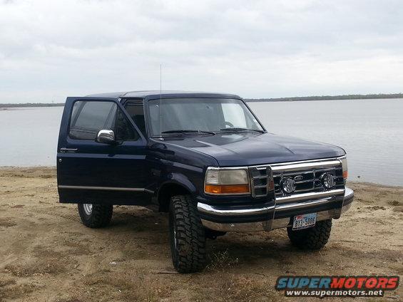 Dark blue SUV with driver's door open, parked on a sandy shore. Chrome grille, aftermarket lights, and off-road tires are visible