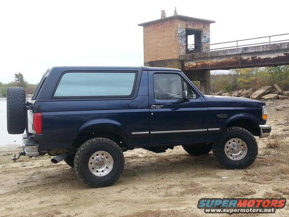 Dark blue Ford Bronco, side view, with spare tire on rear door, parked on dirt near water and a concrete structure