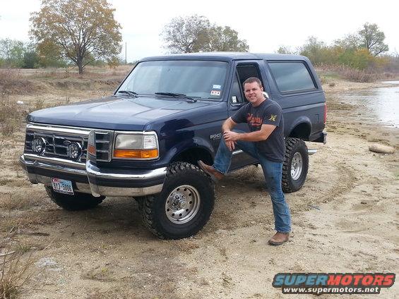 Guy posing with a lifted blue Ford Bronco, possibly early 90s model