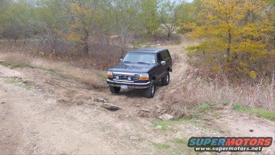 1995 Ford Bronco driving on a dirt trail with trees and dry grass