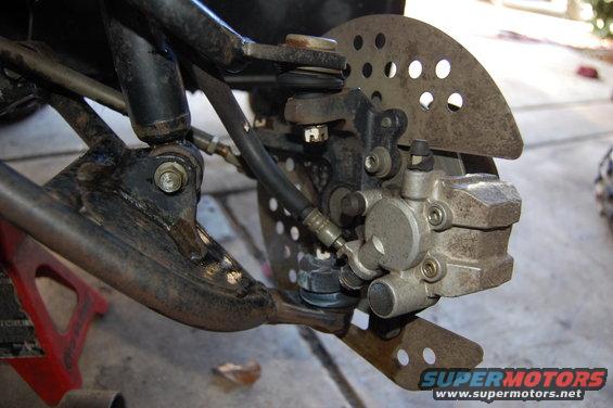 Close-up of ATV front brake caliper, rotor, and suspension components. Supermotors.net logo visible