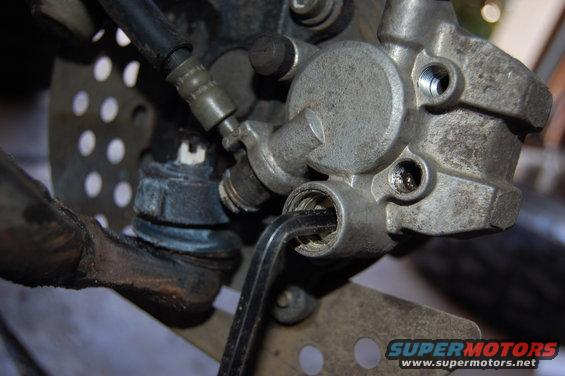 Motorcycle brake caliper with hex wrench inserted, showing perforated disc and suspension components