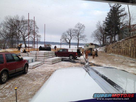 construction site with stacked lumber, a red SUV, a red pickup truck, and a large truck near a body of water
