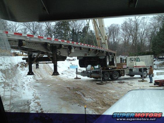 Flatbed trailer with red and white reflectors, supported by jacks, is being loaded onto a flatbed truck with a crane