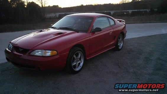 Red Ford Mustang coupe parked on asphalt with trees and train cars in background