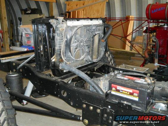 Truck chassis with radiator, fan, and battery installed. Some red machinery is visible in the background