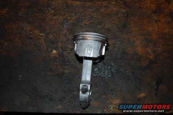 Piston and connecting rod assembly on a wooden surface. The piston has three rings. The connecting rod is attached with two bolts