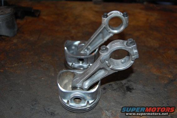Two pistons with connecting rods attached, resting on a wooden surface