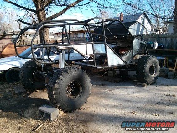 Custom buggy frame with large tires, unfinished metal panels, and roll cage, parked outdoors on concrete