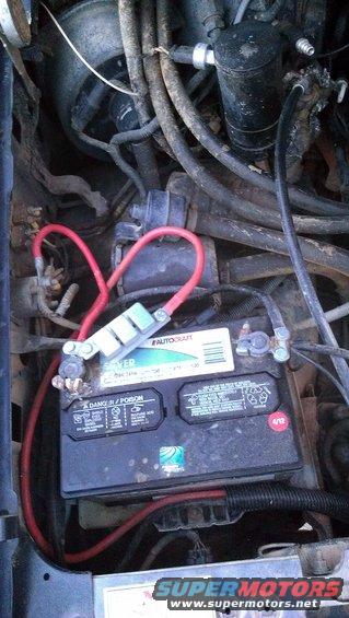 Engine bay showing a car battery with red cables connected. Some hoses and wires are visible
