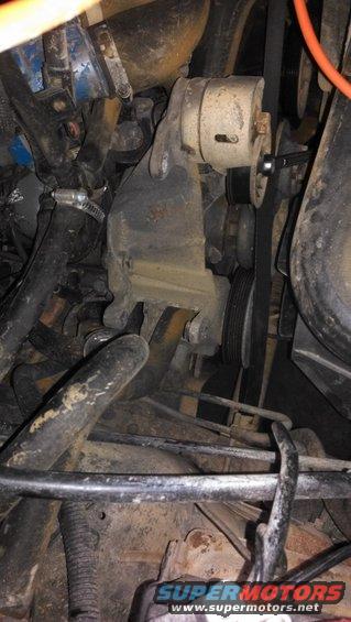 Engine belt tensioner assembly with pulley and mounting bracket, hoses and belts visible